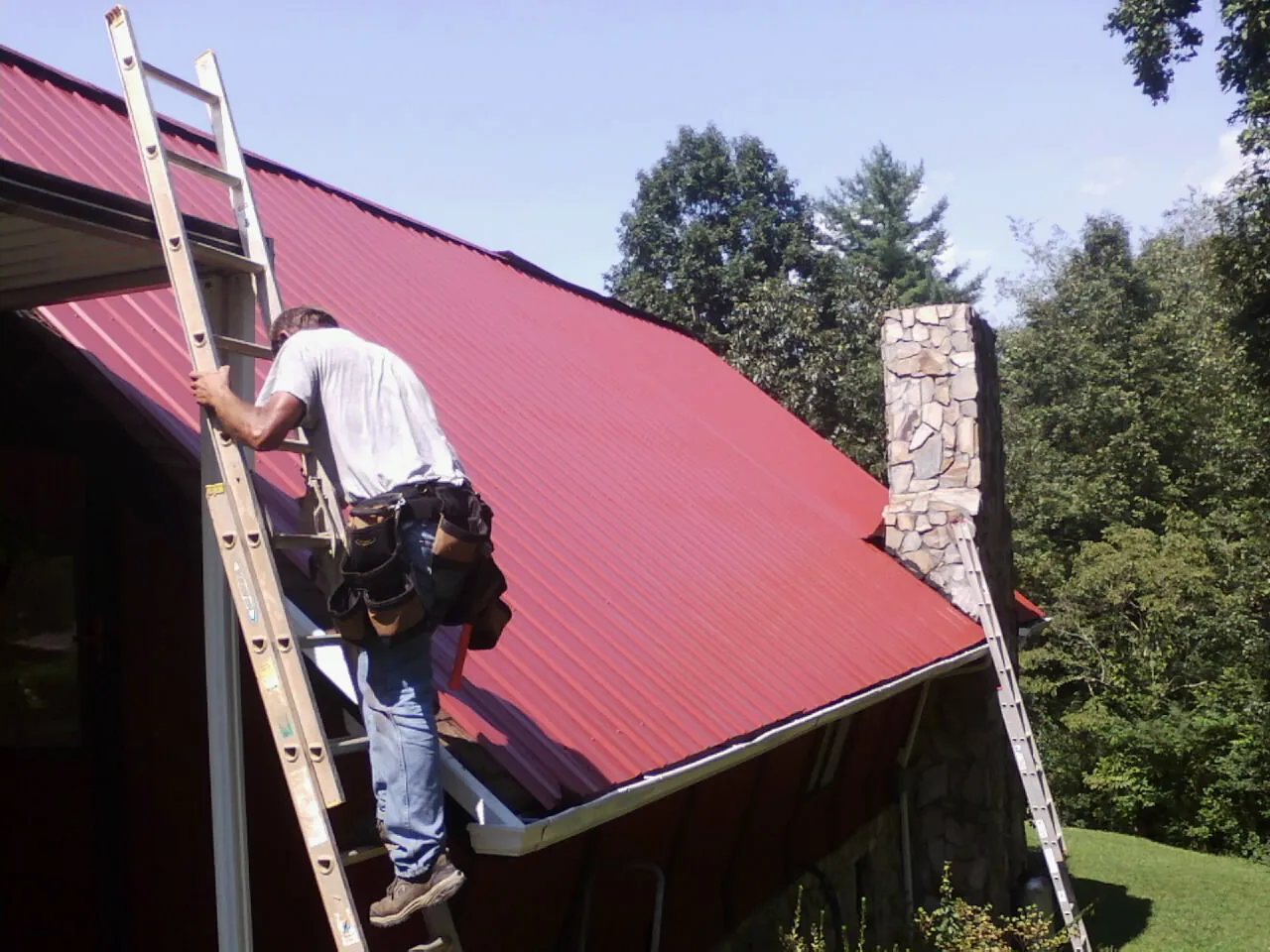 Roofer on a ladder beside a red metal roof and stone chimney — Soffit & Fascia Repair by Roofing Brothers in Gay